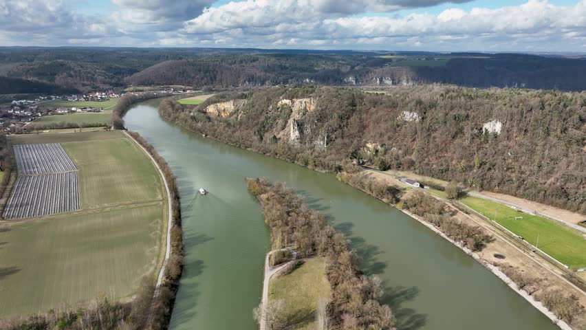 An aerial view of a boat on the Danube River, with a town, agricultural fields, and dramatic rocky cliffs in the background. A great view of the German landscape.