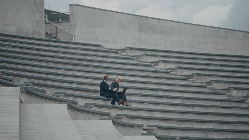 Wide shot of two middle-aged business colleagues sitting on outdoor tiered city stairs while using laptop and having discussion