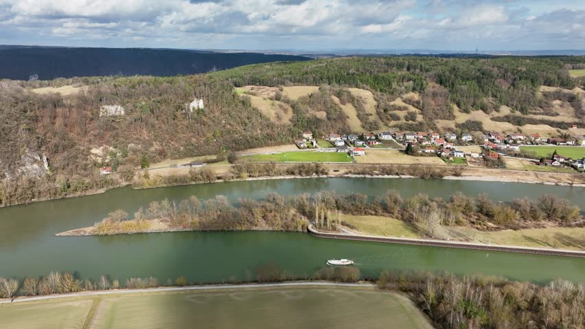 An aerial view of a boat on the Danube River, with a town, agricultural fields, and dramatic rocky cliffs in the background. A great view of the German landscape.
