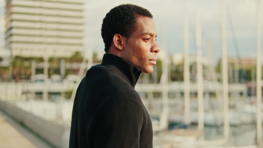 Young man in black turtleneck standing alone by harbor with sailboats and a city in the background, appearing thoughtful.
