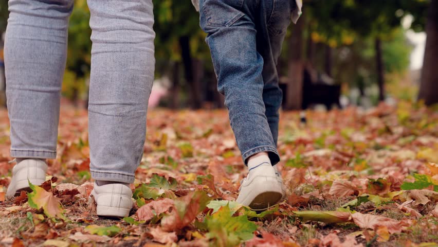 Happy family walk, mom baby in autumn park. Legs, baby mom play together holding hands, autumn outdoors. Happy baby mom walk kicking dry yellow leaves with mom in autumn park. People in nature