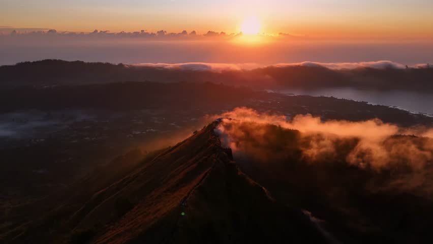 A golden sunrise cast light over Mount Batur in Bali, Indonesia, with hikers gathered at the summit to witness the mist veiling volcanic peak and glowing clouds.