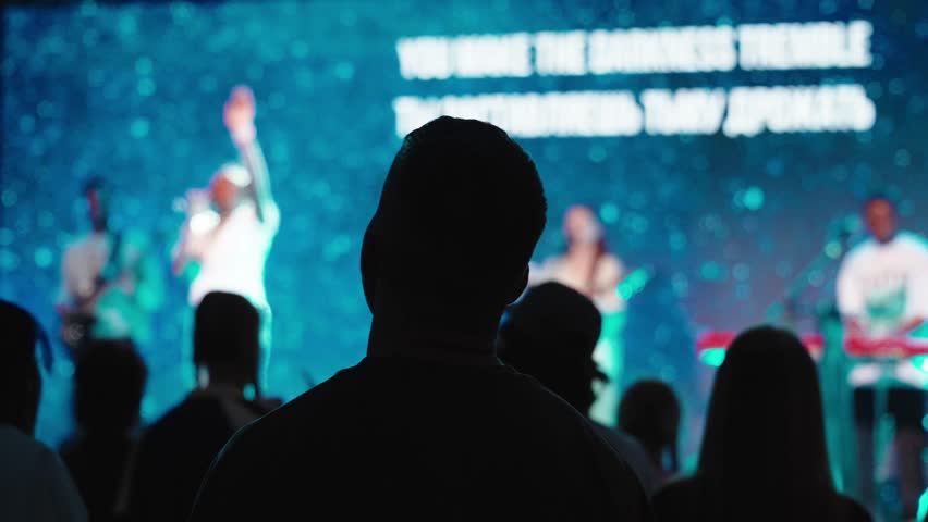 Silhouetted worshipper raising arms during christian concert, standing among crowd, band performing on stage with large screen displaying spiritual messages