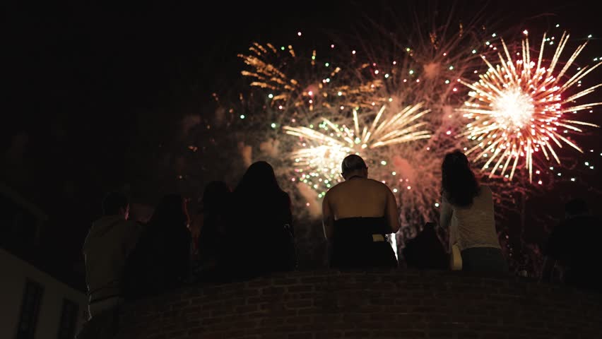 People sitting in the foreground enjoying stunning fireworks in the night sky, festive mood, holiday celebration, New Year’s Eve, fiesta, carnival, dazzling lights, joyful emotions, city fest, slow