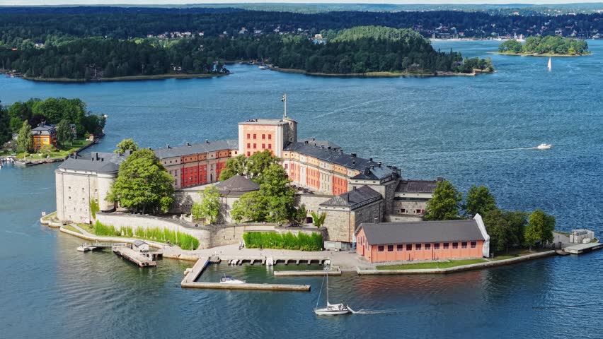 The historic stone Vaxholm Fortress stands surrounded by water and lush greenery, with a Swedish flag flying proudly atop its walls.