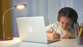 Young girl wearing headphones and sitting at desk with laptop. Young girl is learning how to use laptop in distance. Child is studying at a table during lifestyle evening lessons. A young girl is. - Powered by Shutterstock - Get 15% off with code: PIKWIZARD15