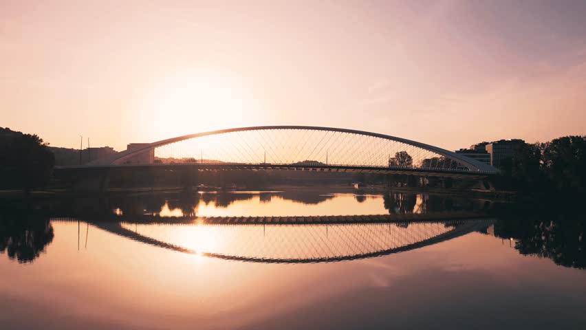 Sunrise casts a golden glow over Troja Bridge as it stretches elegantly across the Vltava River in Prague. The tranquil morning scene reflects beautifully on the water
