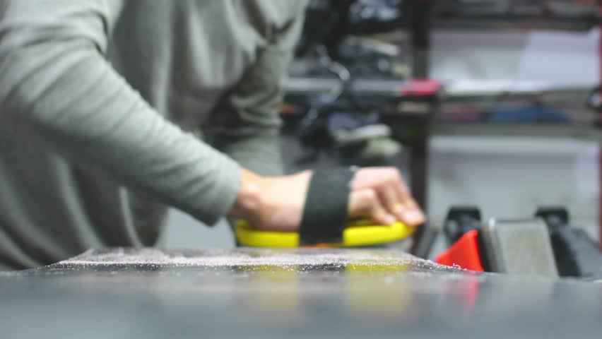 Hand hold brush and brushing wax on snowboard close up. Snowboard preparation in ski repair shop. Intentional blur