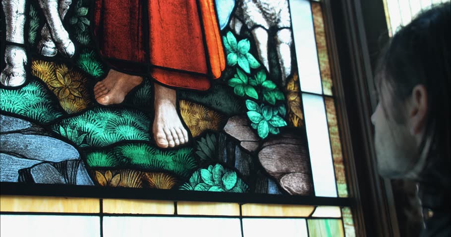 A man stands inside a church, praying at stained-glass window of Jesus the Good Shepherd, evoking themes of spirituality, faith, and quiet reflection.