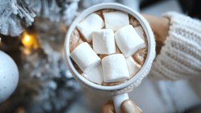 Cozy winter scene features a woman's hands holding a mug of hot chocolate topped with marshmallows, set against a backdrop of a decorated christmas tree with warm, inviting lights - Powered by Shutterstock - Get 15% off with code: PIKWIZARD15