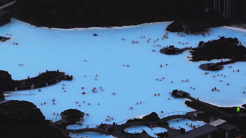Aerial view of the Blue Lagoon Spa in Iceland, with milky blue waters, visitors moving through pools, and steam rising near modern spa buildings.