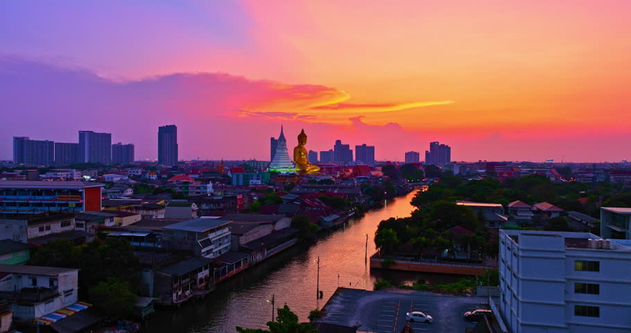 A breathtaking sunset over Bangkok, where the golden Buddha at Wat Paknam Phasi Charoen glows warmly against a dramatic twilight sky, with reflections shimmering across the peaceful canal below.