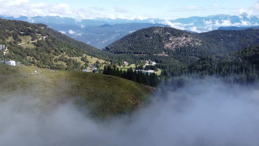 A stunning panoramic view of Soriska planina, showcasing alpine meadows, dense forests, ski lifts, a small lake, and distant mountain ranges under clouds and blue sky.