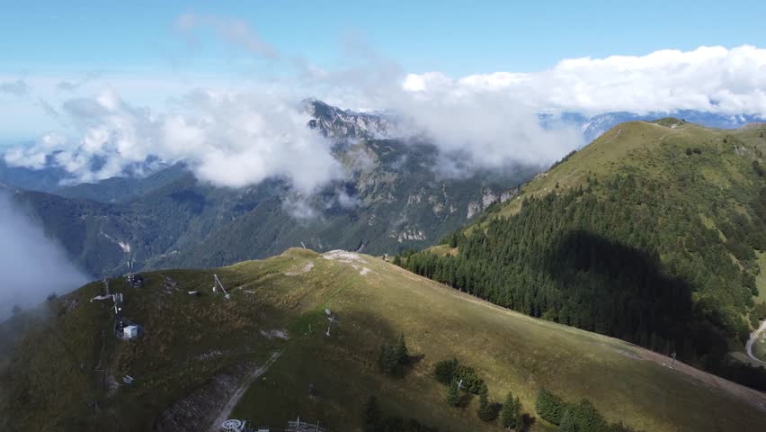 A stunning panoramic view of Soriska planina, showcasing alpine meadows, dense forests, ski lifts, a small lake, and distant mountain ranges under clouds and blue sky.