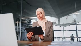 Smiling middle eastern woman in suit and hijab working using touchpad computer. Focused young arabian entrepreneur businesswoman holding digital pc tablet sitting at desk at modern office workplace - Powered by Shutterstock - Get 15% off with code: PIKWIZARD15