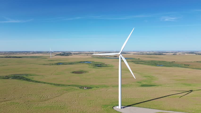 Cinematic aerial drone footage revealing a large, modern wind turbine with spinning blades. The camera starts on a wide, open landscape and pans right to unveil the symbol of green energy.