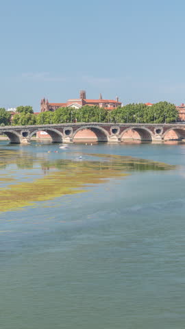 Garonne River and Pont Neuf timelapse in downtown Toulouse, France. Renaissance arch bridge reflects in the water under a blue sky with Basilica of Our Lady of the Daurade. Waterfront with green trees