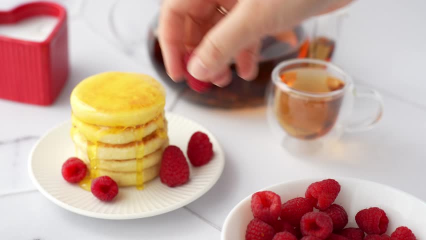 Hand placing fresh raspberry on top of mini pancake stack with honey drizzle and berries served with tea on white table for breakfast. Homemade gourmet breakfast, food styling, culinary presentation.