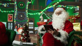 Elderly man dressed as Santa Claus looking at letter received from kids on tablet. Older person wearing Santa costume reads wish lists written by children for Christmas on digital device, camera A - Powered by Shutterstock - Get 15% off with code: PIKWIZARD15