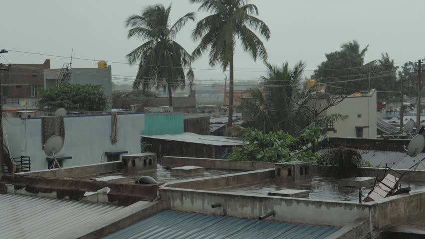 rooftops in a rural or semi-urban Indian town during rainfall. The image captures the moody atmosphere of the monsoon season with visible raindrops, wet roofs, satellite dishes, and tropical