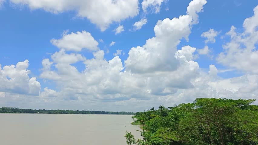 This image captures a calm riverbank with lush greenery under a bright blue sky filled with fluffy white clouds. A perfect portrayal of serene rural nature and peaceful landscape scenery.