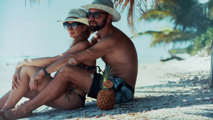 Couple Enjoying Tropical Beach Vacation Relaxing On Sand. Summer Holiday Getaway. Seaside Escape. Pineapple Cocktail.Exotic Destination, Sun Protection, Beach Hats. Serene Ocean View Serene Ocean Trip