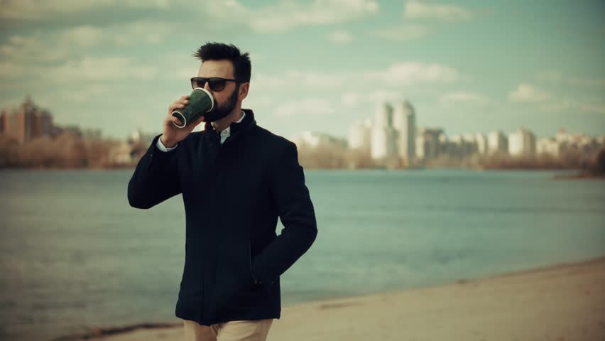 Stylish Man Holding Coffee Cup Walking Along Riverside Promenade. Minimalist Elegance. Leisure, And Serenity. Man Drinking Coffee. Modern Gentleman. Urban Lifestyle Casual Outfit. Calm Trendy Mood