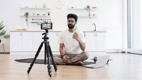 Young adult man recording fitness video in modern home discussing sports and hydration. Includes camera on tripod, yoga mat, water bottle, laptop, white rug, kettle, and minimalistic interior themes. - Powered by Shutterstock - Get 15% off with code: PIKWIZARD15