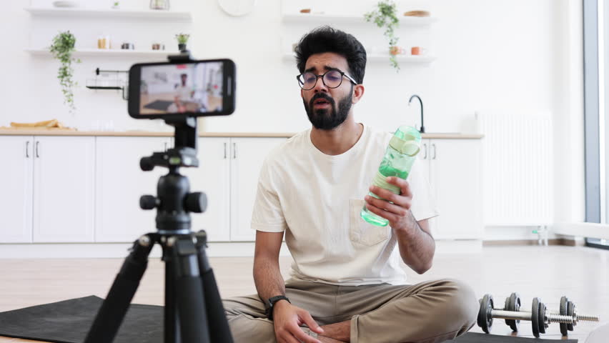 Adult man recording fitness vlog in kitchen holding water bottle explaining hydration benefits during exercise. Young individual speaking confidently to camera promoting healthy living