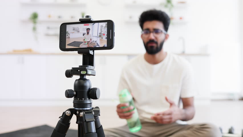 Man in his 30s filming fitness vlog in home kitchen holding water bottle, concept of staying hydrated during workouts, sports, and daily fitness routines, emphasizing health and well-being