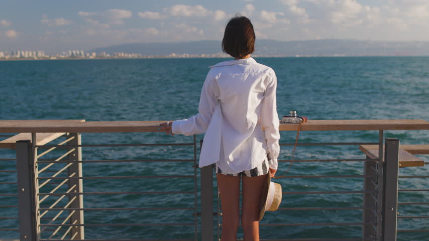 Slender woman in a straw hat looks at the sea and enjoys the moment. Summer vacation by the sea. Beach lifestyle. Freedom and relaxation on the seashore.