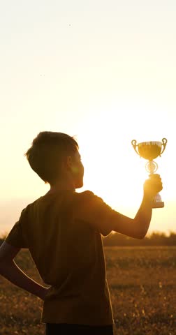 vertical kid hero holds trophy at sunset, celebrating victory in vertical frame. Child kid hero stands proud, glowing sky behind. kid boy success and achievement inspire viewers. Winning motivation