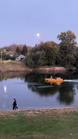 Russia, September 2, 2024:A yellow boat glides on a peaceful river under a full moon, with people enjoying the calm evening. A person walks along the riverbank, adding to the serene landscape