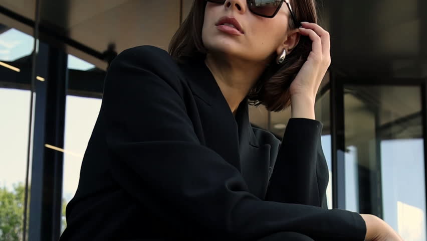 Stylish woman with sunglasses posing against a modern building with large smoky glass windows and concrete walls. Close up of legs walking down steps, wearing leather pants or dress. Fashion ad.