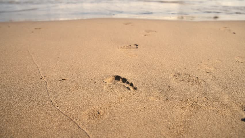Footprints in the sand stretch away from the viewer, leading toward gentle ocean waves on a sunlit beach, creating a peaceful atmosphere of solitude and serene tranquility