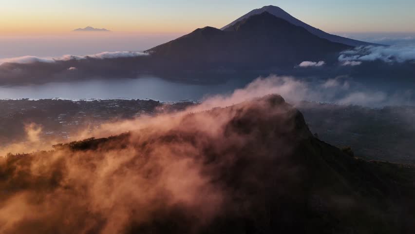 A golden sunrise cast light over Mount Batur in Bali, Indonesia, with hikers gathered at the summit to witness the mist veiling volcanic peak and glowing clouds.