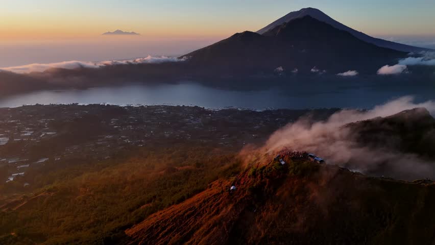 A golden sunrise cast light over Mount Batur in Bali, Indonesia, with hikers gathered at the summit to witness the mist veiling volcanic peak and glowing clouds.