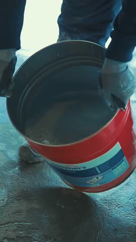 Construction worker pouring gray epoxy resin from a bucket onto a concrete floor