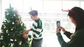 Young adults decorating a Christmas tree at home near bright windows, smiling as one takes a smartphone photo of the other; a festive holiday scene with ornaments and lights. 4k video - Powered by Shutterstock - Get 15% off with code: PIKWIZARD15