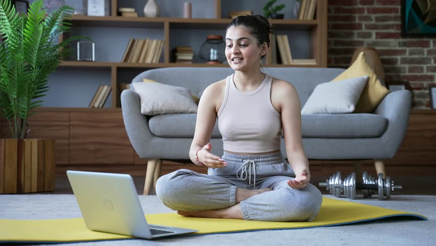 Confident female yoga trainer giving video call lesson using laptop sitting on mat on floor in living room at home. Young woman coach has online course engages in remote training or meditation session