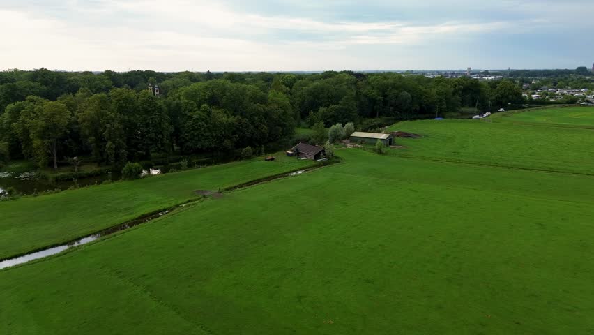 A winding stream flows through wide green meadows, with small rustic barns near the forest edge and a distant town under a calm, cloudy sky.