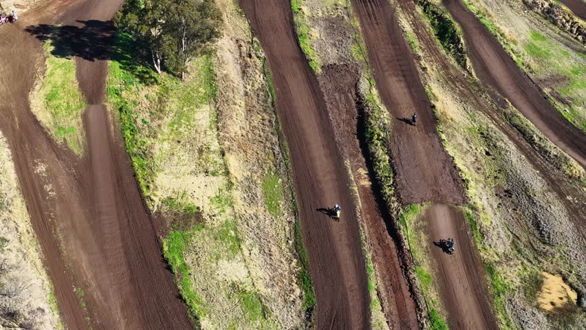 Multiple motocross riders speed along winding dirt track, captured in bright daylight from above