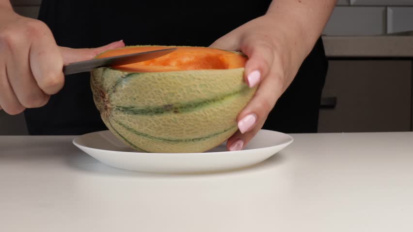 Cutting orange melon into slices on white plate. Close-up of hands preparing fresh cantaloupe fruit in home kitchen for healthy snack or meal prep