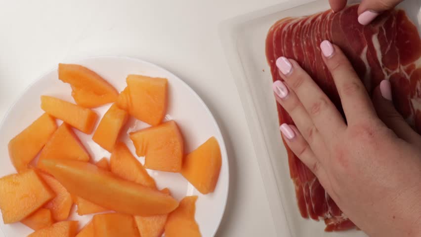 Wrapping slices of melon with jamon on white plate. Hands preparing prosciutto and fruit appetizer in home kitchen for party or gourmet snack