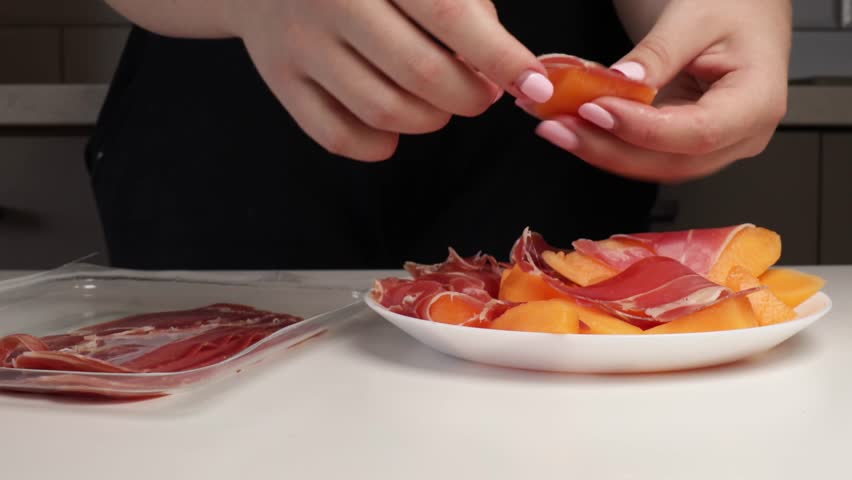 Wrapping prosciutto around melon slices on white plate. Hands preparing traditional appetizer in home kitchen for serving or entertaining