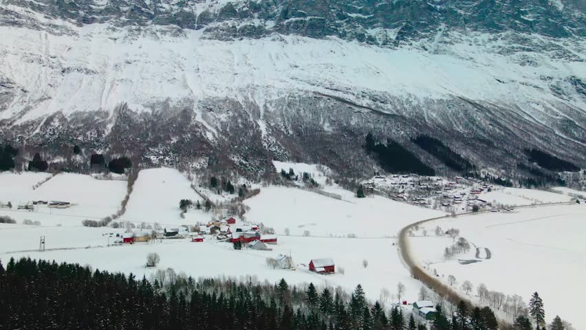 Drone footage captures the winter landscape of Geirangerfjord, Norway. The iconic fjord is surrounded by snow covered mountains, with a charming village nestled in the valley. Perfect for travel, natu