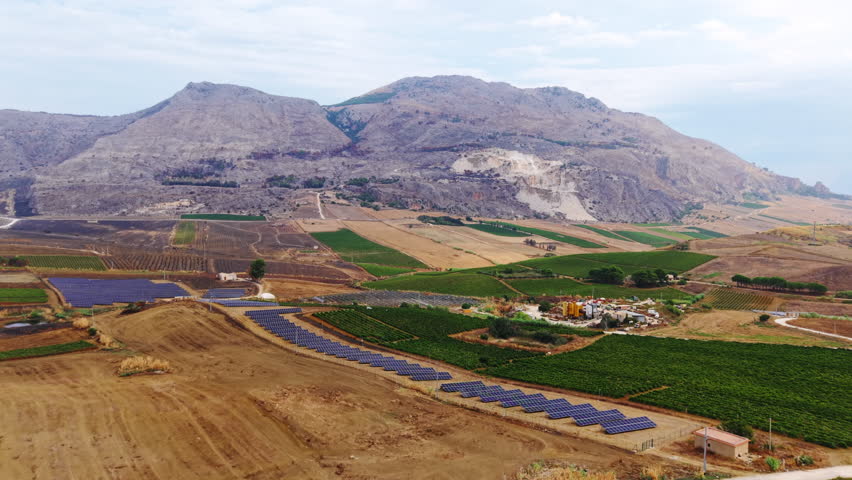 Wide drone shot in Sicily showing solar panel fields, dry soil, cultivated land, water pond, scattered houses, roads, and distant mountains.