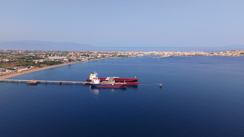 Drone shot approaching oil tanker docked near offshore refinery with Sicilian coastline and bay in the background