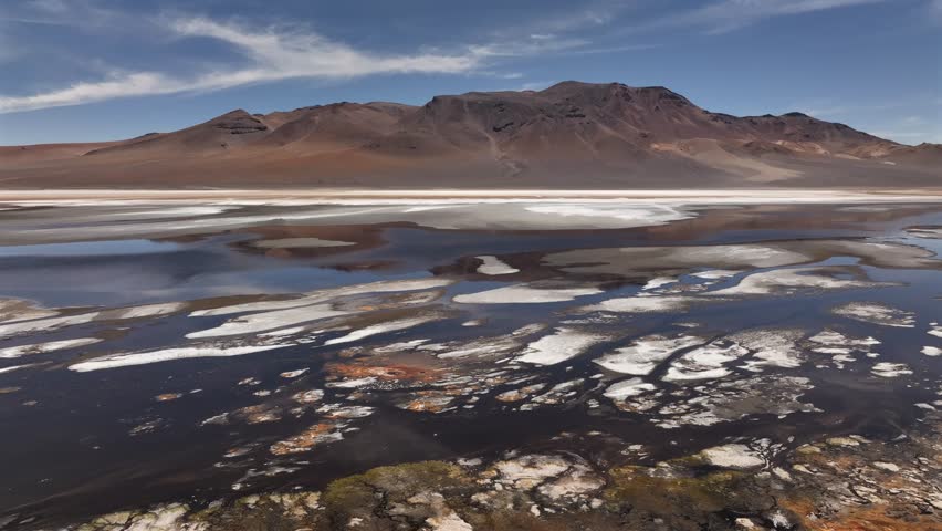 Aerial view of the rugged Ruta 27 landscape meeting the serene Salar y Laguna de Tara, contrasting with the distant mountain range, Salar y Laguna de Tara, Antofagasta, Chile.