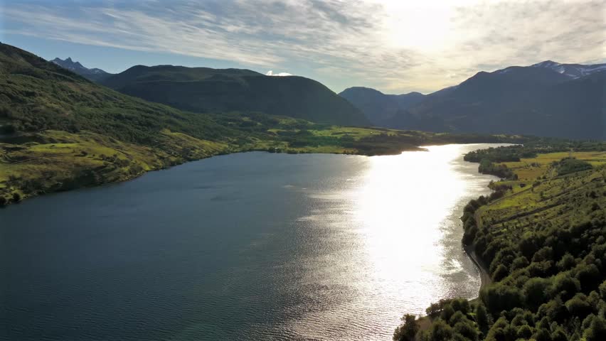 Aerial view of dark blue lake reflecting sunlight, bordered by verdant green hills and majestic mountains under a bright sky, Coyhaique, Aysén, Chile.